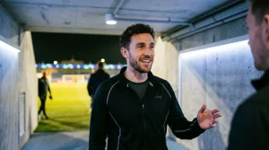 Mann in schwarzer Sportjacke im Stadiongang lächelnd